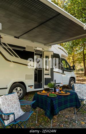 Casa a motore in campeggio con un tavolo e sedie allestiti all'esterno del veicolo e pronti a mangiare e bere. Foto Stock