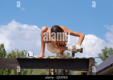 Un falegname lavora con un cacciavite elettrico giallo, ruota una vite in una trave di legno della tavola sul tetto Foto Stock