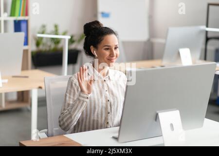 Riunione online. Donna allegra che fa videochiamata sul computer in ufficio, lavoratore di ufficio femminile in airpod che ondano alla webcam Foto Stock