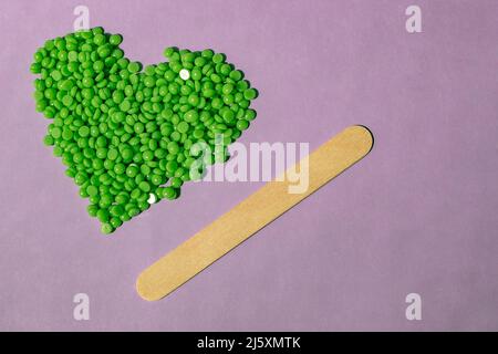 Bellissimi granuli di cera verde con estratto di aloe per la depilazione sono versati su uno sfondo viola chiaro, a forma di cuore. spatola di legno. Epilat Foto Stock