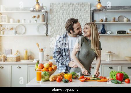 Soddisfatto giovane europeo maschio con hugs stoppie femmina, prepara il cibo a tavola con verdure biologiche in cucina Foto Stock