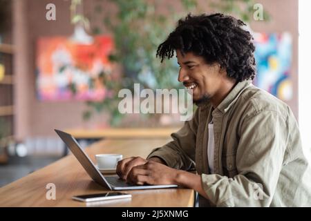 Allegro giovane africano americano curly maschio digitando sul portatile a tavola con caffè e telefono in interno caffè Foto Stock