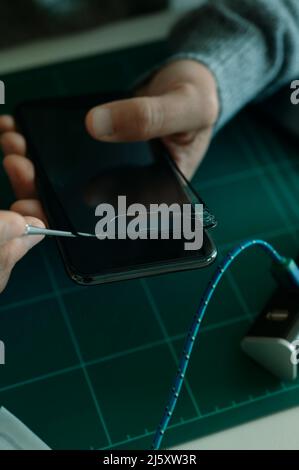 un giovane uomo caucasico rimuove il proteggischermo rotto di uno smartphone, seduto su un piano di lavoro Foto Stock