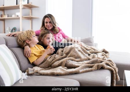 Mamma allegra con bambini simpatici che guardano un film interessante mentre riposano insieme su un comodo divano in un soggiorno luminoso a casa Foto Stock