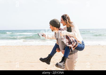 Vista laterale dell'uomo che si prende cura di dare giro piggyback alla ragazza allegra sulla costa sabbiosa mentre prende selfie in giorno di sole Foto Stock