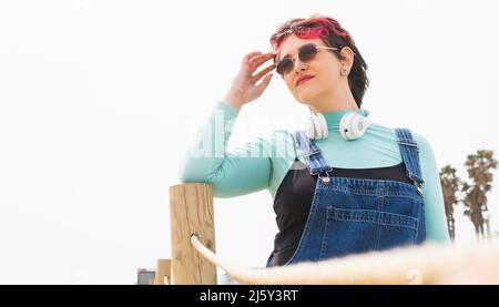 Donna con capelli corti colorati in occhiali da sole e cuffie sul collo guardando lontano mentre tocca la fronte vicino ringhiera di legno Foto Stock