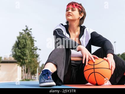 Donna in abbigliamento sportivo con capelli corti tinti seduti su campo sportivo con basket e guardando via Foto Stock