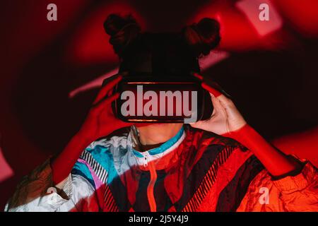 Giovane donna in visore VR che esplora la realtà virtuale mentre si alza su sfondo rosso in studio con luce soffusa Foto Stock