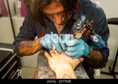 uomo in lattice guanti che fanno tatuaggio su dito di irriconoscibile cliente con macchina speciale in salone Foto Stock