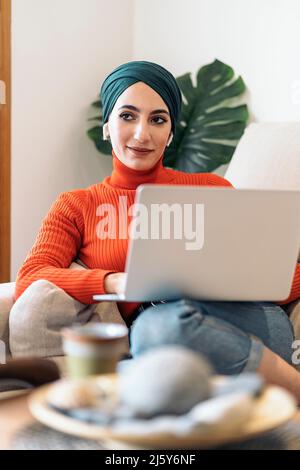Femmina musulmana in elegante dolcevita e foulard sorridente e usando il laptop mentre ascolti la musica sul divano in giornata di sole a casa Foto Stock