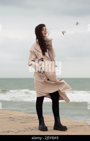 Corpo pieno di donna in cappotto in piedi guardando lontano vicino mare ondeggiante sulla costa sabbiosa con gabbiano bianco volante in tempo ventoso Foto Stock