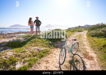 Coppia in piedi sulla spiaggia di sabbia soleggiata dell'oceano con la bicicletta Foto Stock