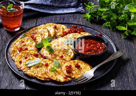 frittelle dolci di farine di farro con salsa di bacca, mirtillo di montagna, cowberry su piatto nero su tavola di legno scuro, vista orizzontale dall'alto Foto Stock