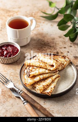 Crepes in piastra marrone. Composizione della colazione con blinis, forchette rustiche, tè, marmellata e foglie verdi sul retro. Orientamento verticale Foto Stock