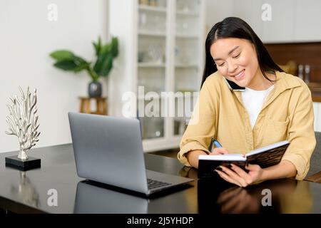 La donna freelance asiatica parla sullo smartphone e prende appunti con la penna, utilizzando un computer portatile per il lavoro remoto da casa, scrivere alcune informazioni seduti al tavolo. Concetto di multitasking Foto Stock
