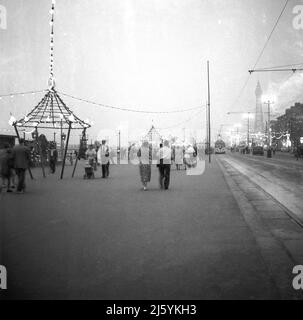 1960, storico, prima sera e la gente passeggiare lungo il fronte a Blackpool, Inghilterra, Regno Unito. La linea del tram si trova sulla destra del lungomare, mentre la famosa torre della città è visibile in lontananza. Foto Stock