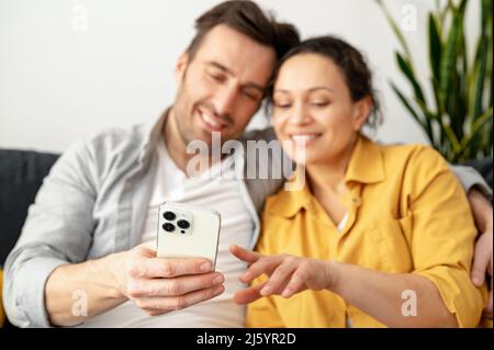 Coppia caucasica eccitata e gioiosa seduta sul divano a casa e con smartphone in mano. Uomo e donna che guardano film o leggono messaggi Foto Stock