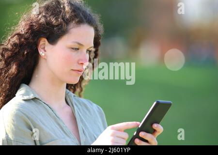 Donna seria che usa lo smart phone che cammina in un parco una giornata di sole Foto Stock