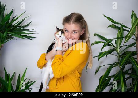 giovane donna bionda che porta il gatto bianco calico sulle braccia abbracciando il kitty sorridendo alla macchina fotografica guardando felice con lo spazio di copia Foto Stock