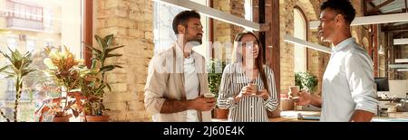 Bello parlare. Tre giovani colleghi in abbigliamento casual holding tazze da caffè e discutere di qualcosa di permanente, mentre nell'ufficio moderno Foto Stock
