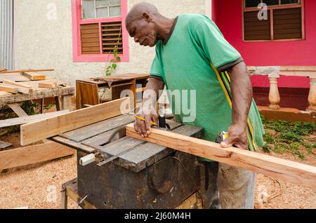 Un costruttore di cabinate con un pennel tra le sue dita sostiene un pezzo di legno 2 x 2 come usa una vecchia sega elettrica per tagliare la sua estremità Foto Stock