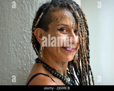 Attraente alternativa giovane messicana donna con lunghissimo temibile Rasta trecce, body art viso piercing e tatuaggio sembra positivamente ottimista al telespettatore. Foto Stock