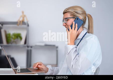 Il medico si consulta al telefono e utilizza un computer portatile in ufficio Foto Stock