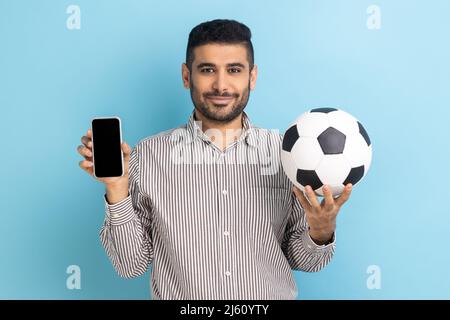 Sorridente uomo bearded soddisfatto che tiene la sfera di calcio e lo smartphone vuoto nero display, prenotazione biglietti per il campionato, indossando maglia a righe. Studio interno girato isolato su sfondo blu. Foto Stock