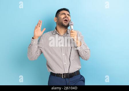 Ritratto di bel giovane uomo adulto con barba che canta le canzoni preferite con microfono in mano, indossando una camicia a righe. Studio interno girato isolato su sfondo blu. Foto Stock