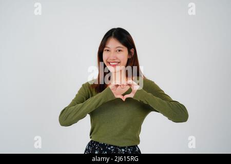 Attraente giovane donna asiatica si sente felice e forme romantiche cuore gesto esprime sentimenti teneri indossa casual jumper pose contro bianco Foto Stock