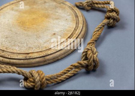 Tavola rotonda di taglio in legno e corda della nave sullo sfondo grigio. Svuotare il vecchio tagliere. Corda doppia di iuta con nodi semplici legati. Vista angolata Foto Stock