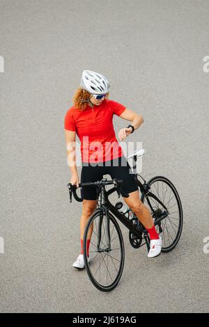 Donna orientata in piedi su un pavimento in cemento con una bicicletta tra le sue gambe. Sta controllando il suo orologio. Foto Stock