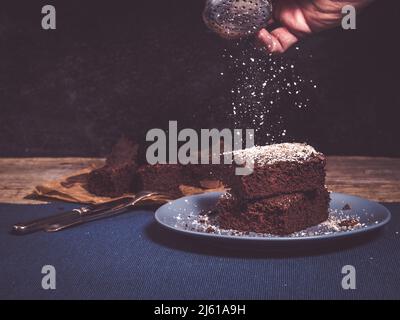 l'immagine mostra brownie appena sfornate su tavola rustica di legno; mano che tiene un dredger shaker; zucchero in polvere sta volando sopra torta di brownie Foto Stock