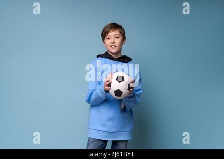 Ragazzo sorridente in abiti blu sport in una felpa che tiene palla di calcio in posa in studio su uno sfondo colorato. Teenager caucasico fan di giocare a calcio Foto Stock