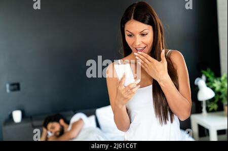 Donna che parla privatamente sul cellulare mentre suo marito dorme sopra letto Foto Stock