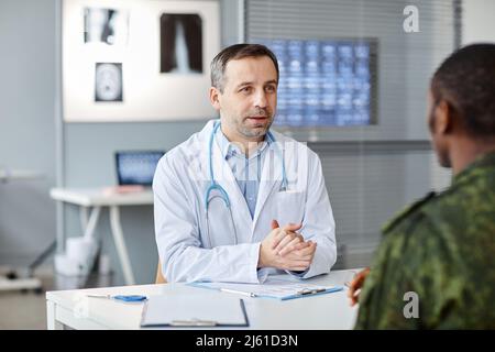 Medico caucasico maturo seduto al banco di fronte al soldato nero irriconoscibile che parla dei test necessari per il controllo della salute Foto Stock