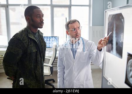 Medico maturo serio che mostra immagine radiologica della colonna lombare al giovane ufficiale militare adulto durante la consultazione in ospedale Foto Stock