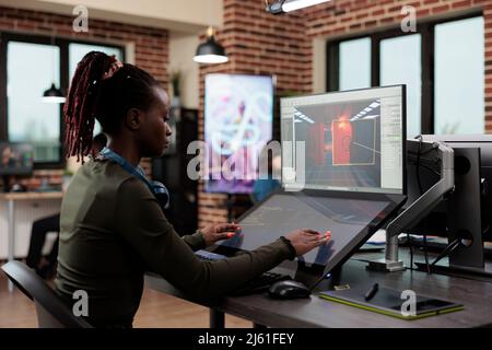 Dipendente dell'azienda di arti elettroniche che lavora al progetto 3D durante la modellazione dell'ambiente. Sviluppatore afroamericano seduto in ufficio agenzia creativa mentre progetta il livello di gioco. Foto Stock