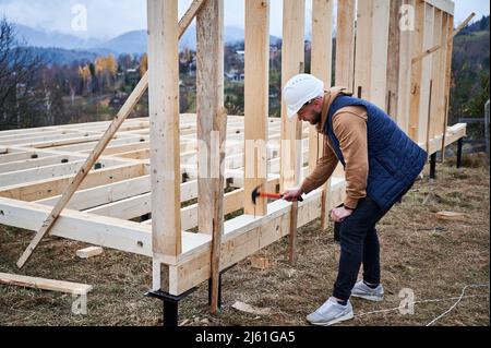 Uomo lavoratore costruire casa di legno telaio su palo fondazione. Il carpentiere martella il chiodo in capriate di legno, usando il martello. Concetto di carpenteria. Foto Stock