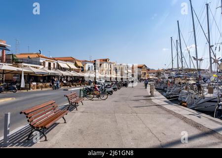 Isola AEGINA, Grecia - 04.27.2022: Persone che camminano nella strada principale con caffè su un lato e il porto e il porto di fronte al pittoresco villaggio Foto Stock