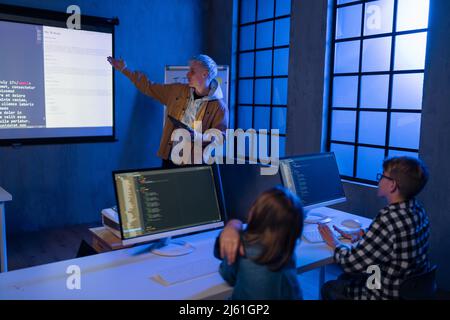 Insegnante che dà lezioni di informatica ai giovani studenti in camera oscura. Camera. Spiegazione della tecnologia dell'informazione. Foto Stock