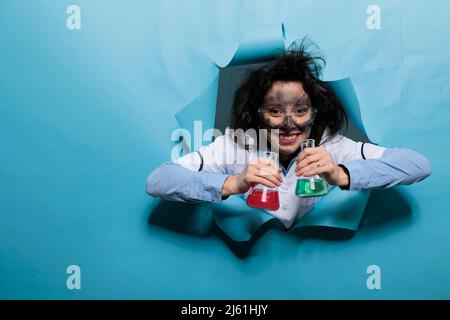 Crazy Silly chemist con volto sporco e capelli wacky che tiene becher di vetro riempito di sostanza liquida mentre su sfondo blu. Scienziato di MAD con l'aspetto divertente che grining terribile alla macchina fotografica. Foto Stock