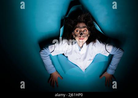 Pazzo esperto di biochimica con l'espressione del viso goofy e l'aspetto wacky che agisce divertente mentre su sfondo blu. Chimico lunatico con capelli disordinati un volto sporco guardando la fotocamera. Foto studio. Foto Stock