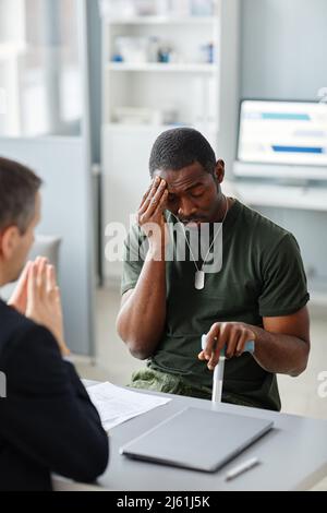 Giovane soldato afroamericano con bastone a piedi seduto di fronte a psicoterapeuta lamentando mal di testa cronico stress Foto Stock