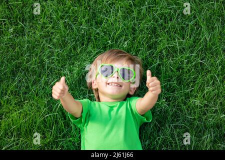 Bambino felice che gioca all'aperto nel parco primaverile. Bambino sdraiato su erba verde. Giornata della terra e concetto di ecologia. Ragazzo che mostra i pollici in su Foto Stock