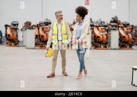 Ingegnere che tiene hardhat a piedi con donna d'affari in fabbrica Foto Stock