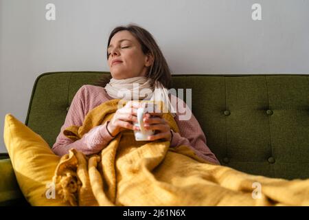 Donna malata che tiene una tazza di tè che riposa sul divano a casa Foto Stock