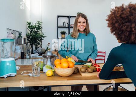 Donna bionda sorridente che taglia le mele con un amico seduto al tavolo in cucina Foto Stock