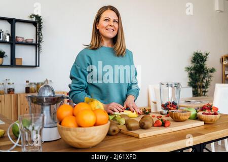 Donna bionda sorridente con frutta fresca al tavolo in cucina Foto Stock
