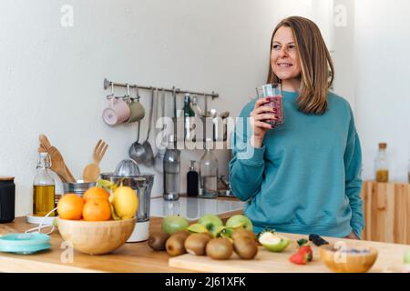 Donna bionda sorridente con bicchiere di frullato in piedi al tavolo in cucina Foto Stock